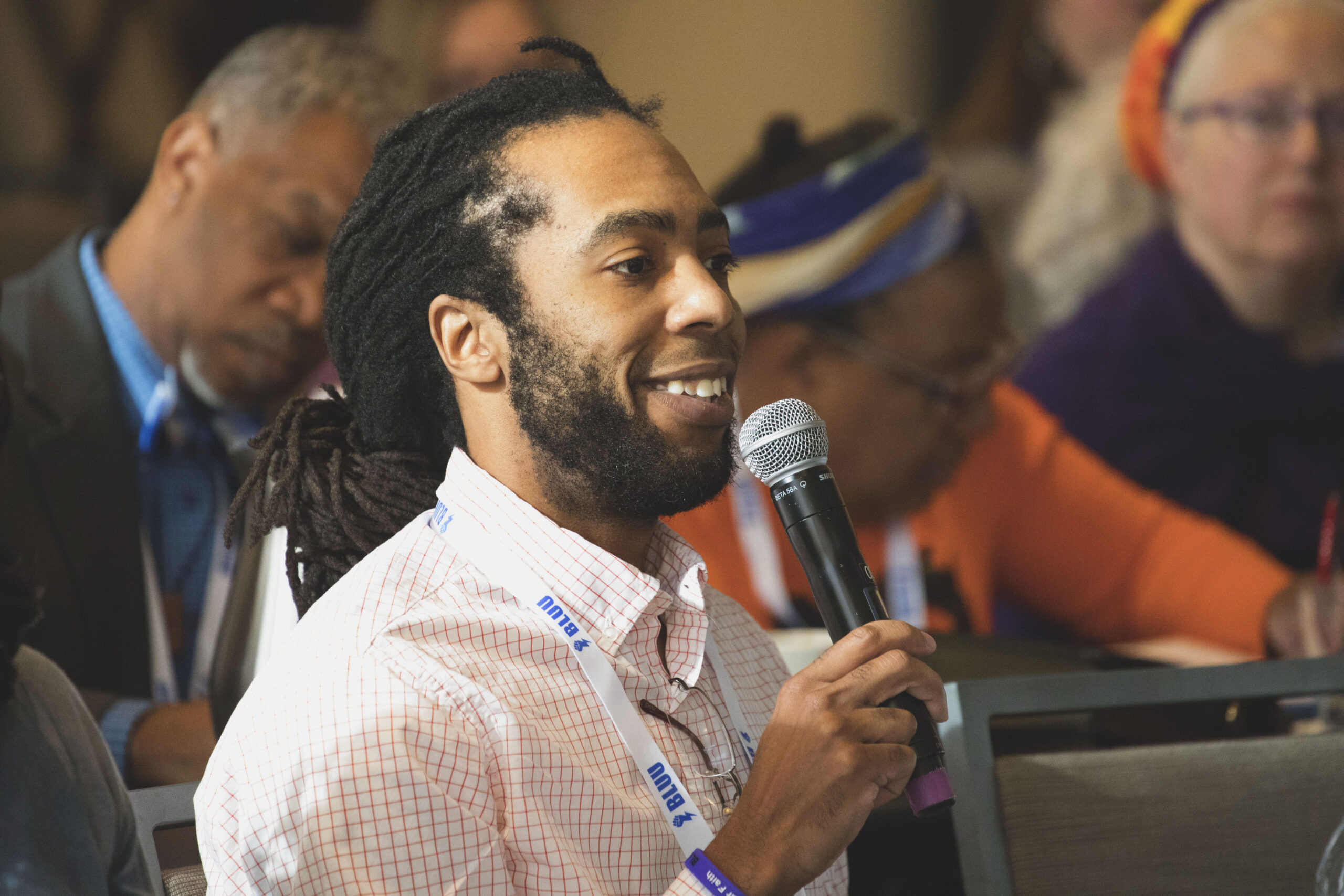 A Black man with locs holds a microphone while smiling.