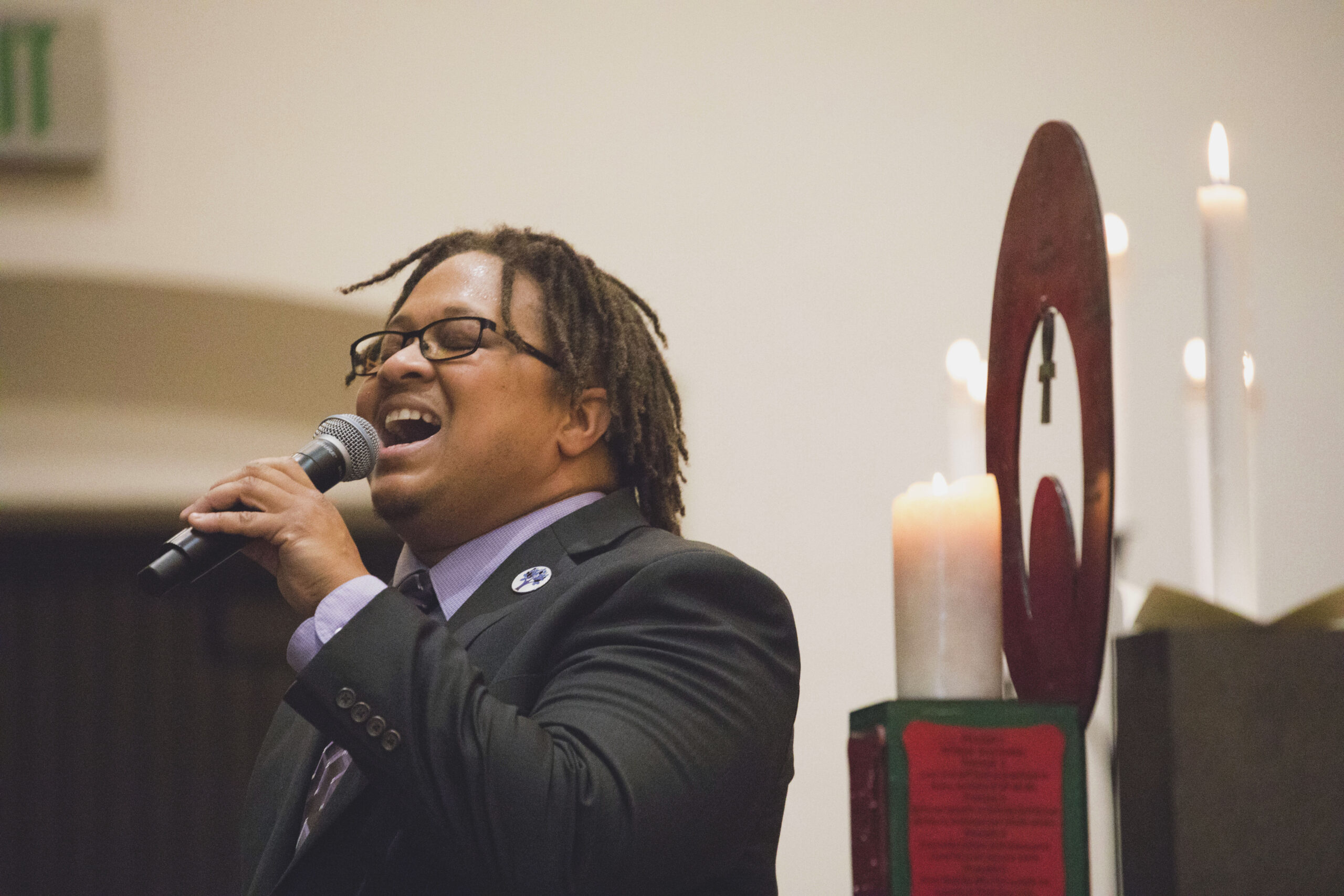 A Black man wearing a suit holds a microphone and sings. He stands in front of candles and a chalice.