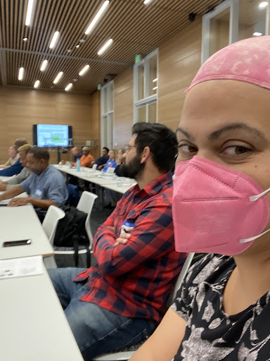 A self of BLUU executive director Lena K. Gardner. She is sitting in a large conference room with a lot of people behind her. In the photo, she is wearing a mask and a bandana.