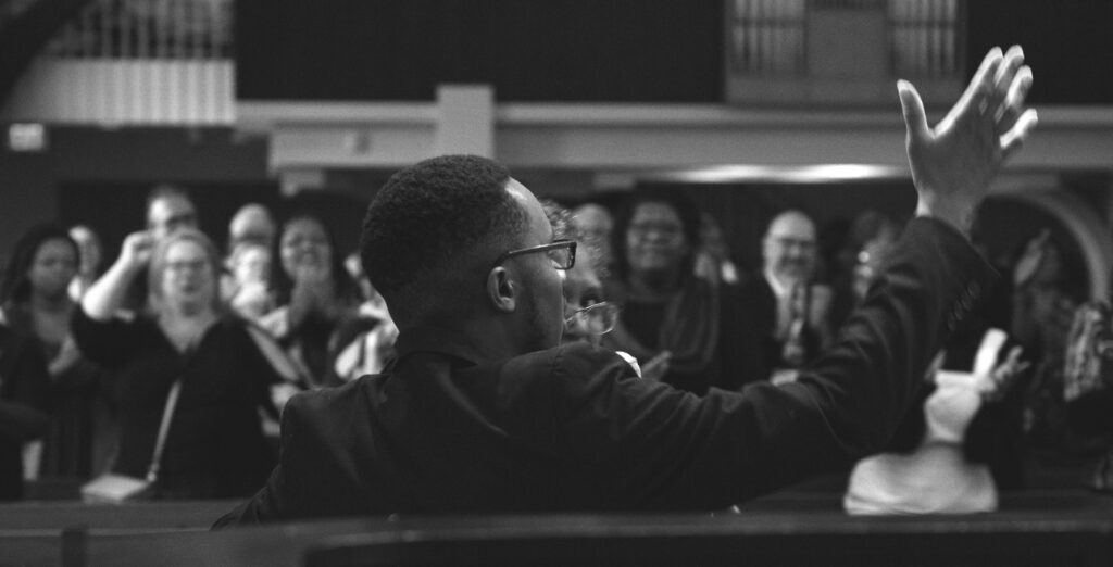 A Black musician, with a fade and glasses, stands with his back facing a crowd. One hair is in the air as if he is conducting the crowd in a song. The crowd's faces are blurred.
