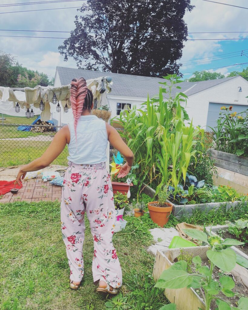 Jessa Rose, a Black woman with a long pink braid, a tank top and floral pants, walks toward a garden.