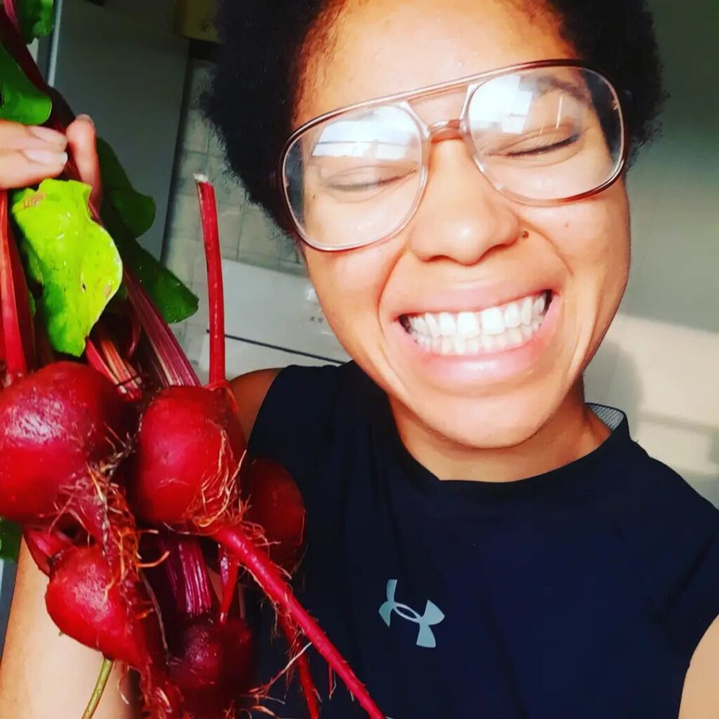 Jessa Rose, a Black woman smiling at the camera and wearing a dark t-shirt and glasses, is holding a handful of beets.