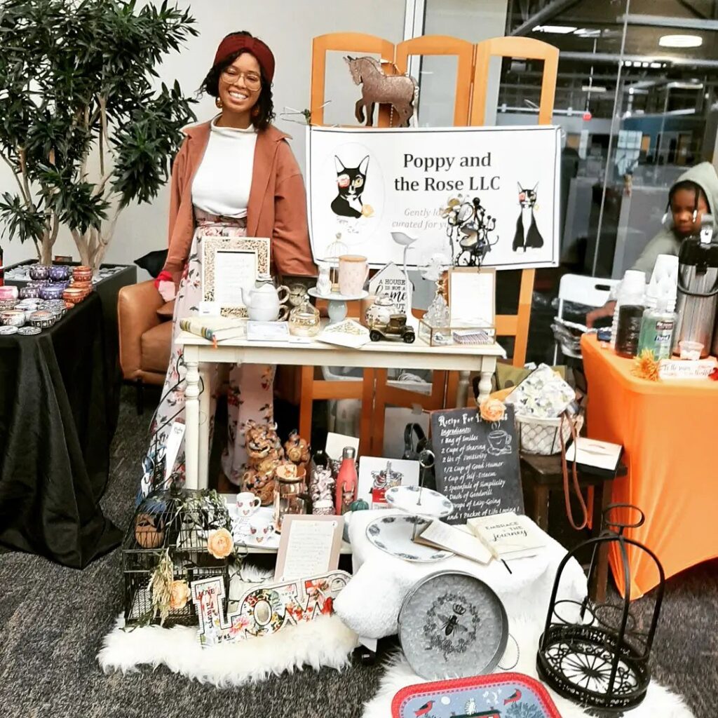 Jessa Rose, a Black woman wearing a headband, glasses, and a top and jacket, stands next to a an assortment of items from her thrifstore.