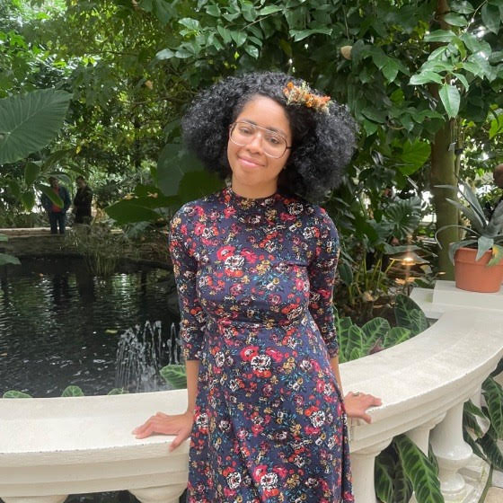 Description: Jessa Rose, a Black woman wearing glasses and a floral dress, stands outside in front of many plants.