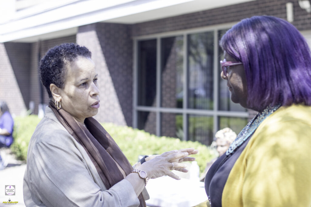A Black woman with short hair wearing a scarf talks to another Black woman with bright hair and wearing glasses. They are standing outside in front of a building.