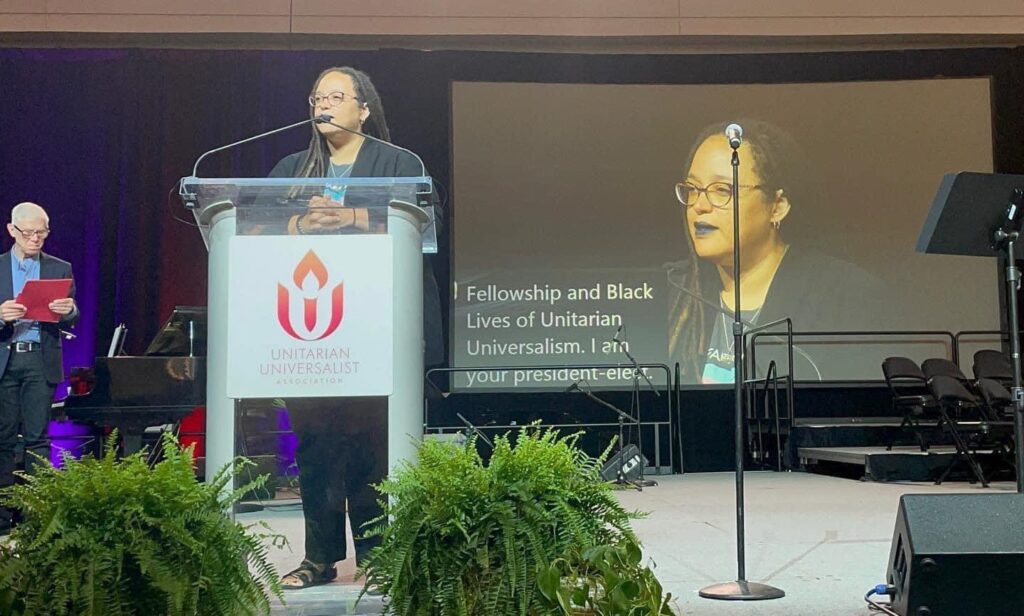 Rev. Dr. Sofia Betancourt, a Black woman with long locks, wearing dark lipstick a cardigan and top, and dark pants, and sandals, stands at a podium with the Unitarian Universalist Association logo on the front.