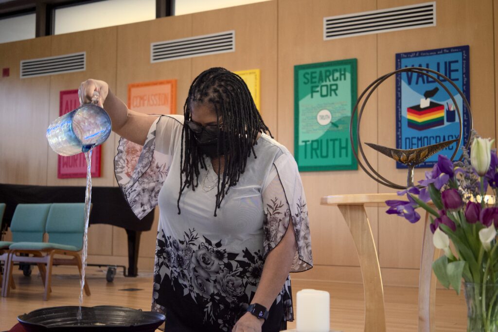 A Black person with locs standing in a church pours a pitcher of water into a receptacle as part of worship.