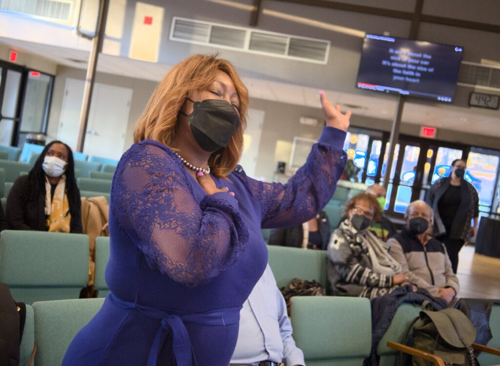 A Black person with red hair  and wearing a floral dress lifts her hand during BLUU worship
