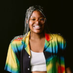 Beatrice Ogeh, a Black person, wears bright clothing while standing in front of a dark background.